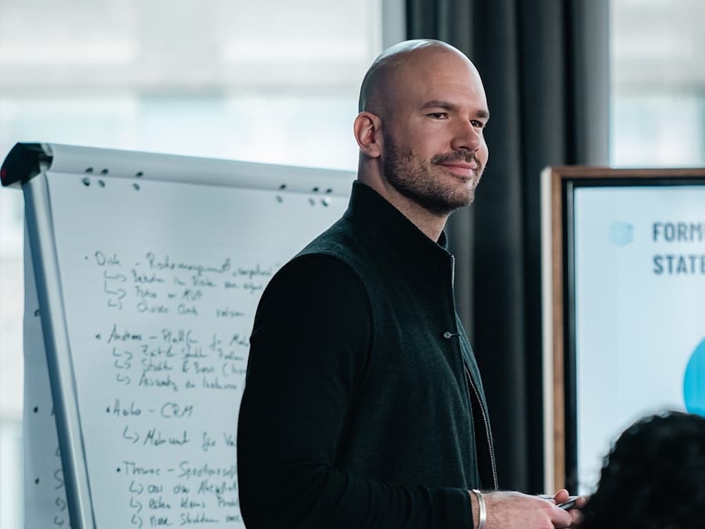 Mann mit Glatze und Bart in schwarzem Pullover steht neben einem Flipchart mit handschriftlichen Notizen und einem Bildschirm im Hintergrund.
